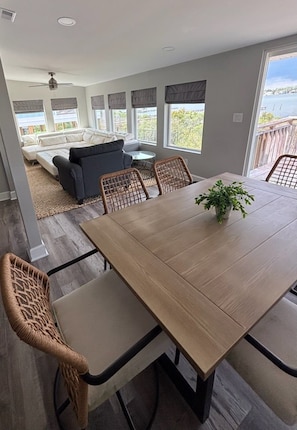 A dining area with a wooden table and wicker chairs, a gray sofa, and a view of a body of water through large windows. - Bay-view 5BR Gulf Shores home with 5.5 baths, sleeps 12, and easy beach access. (Gulf Shores)