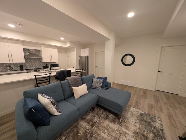 A modern living room with a sectional sofa, a kitchen area with white cabinets, and a dining table.