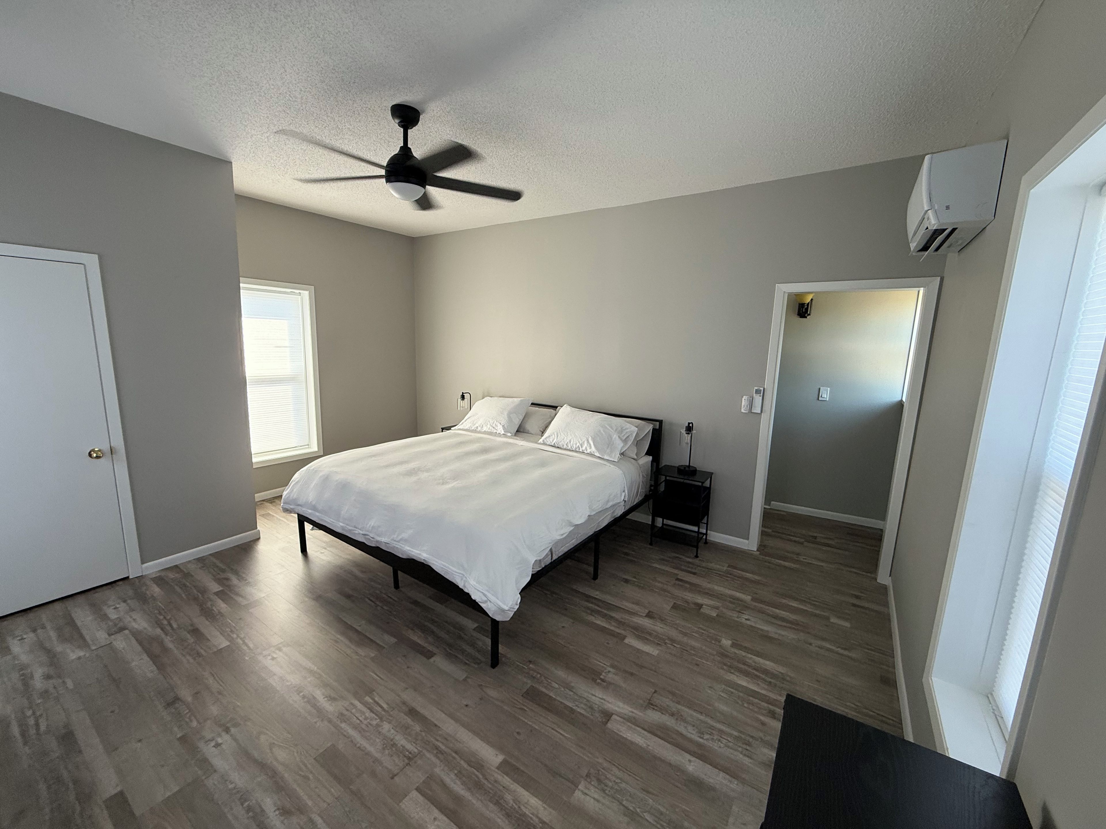 Un dormitorio con cama, ventilador de techo, mesita de noche, aire acondicionado instalado en la pared y una puerta que da a otra habitación.