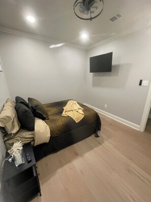A bedroom with a bed, a TV mounted on the wall, a bedside table with a remote, and a ceiling fan.