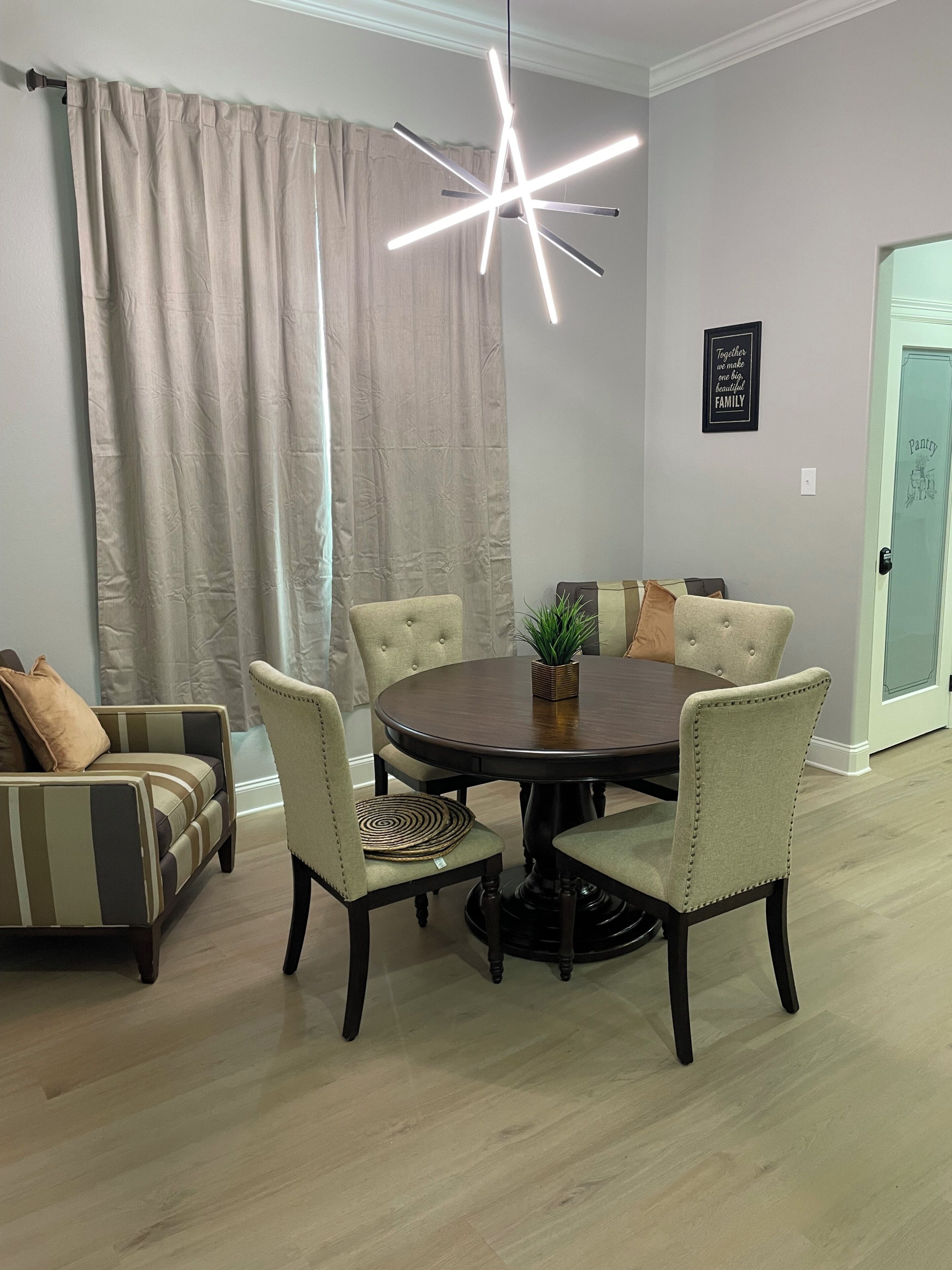 A dining area with a round table, four chairs, a hanging light fixture, a potted plant, and a wall with a framed quote.