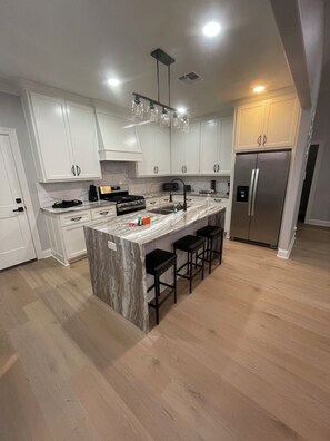 A modern kitchen with white cabinets, a marble island, black bar stools, and stainless steel appliances.