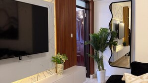 Ein modernes Wohnzimmer mit Flachbildfernseher an einer Marmorwand, einer Wand aus Holzpaneelen, einem großen Spiegel, einer schwarzen Couch und einem Blumentopf.