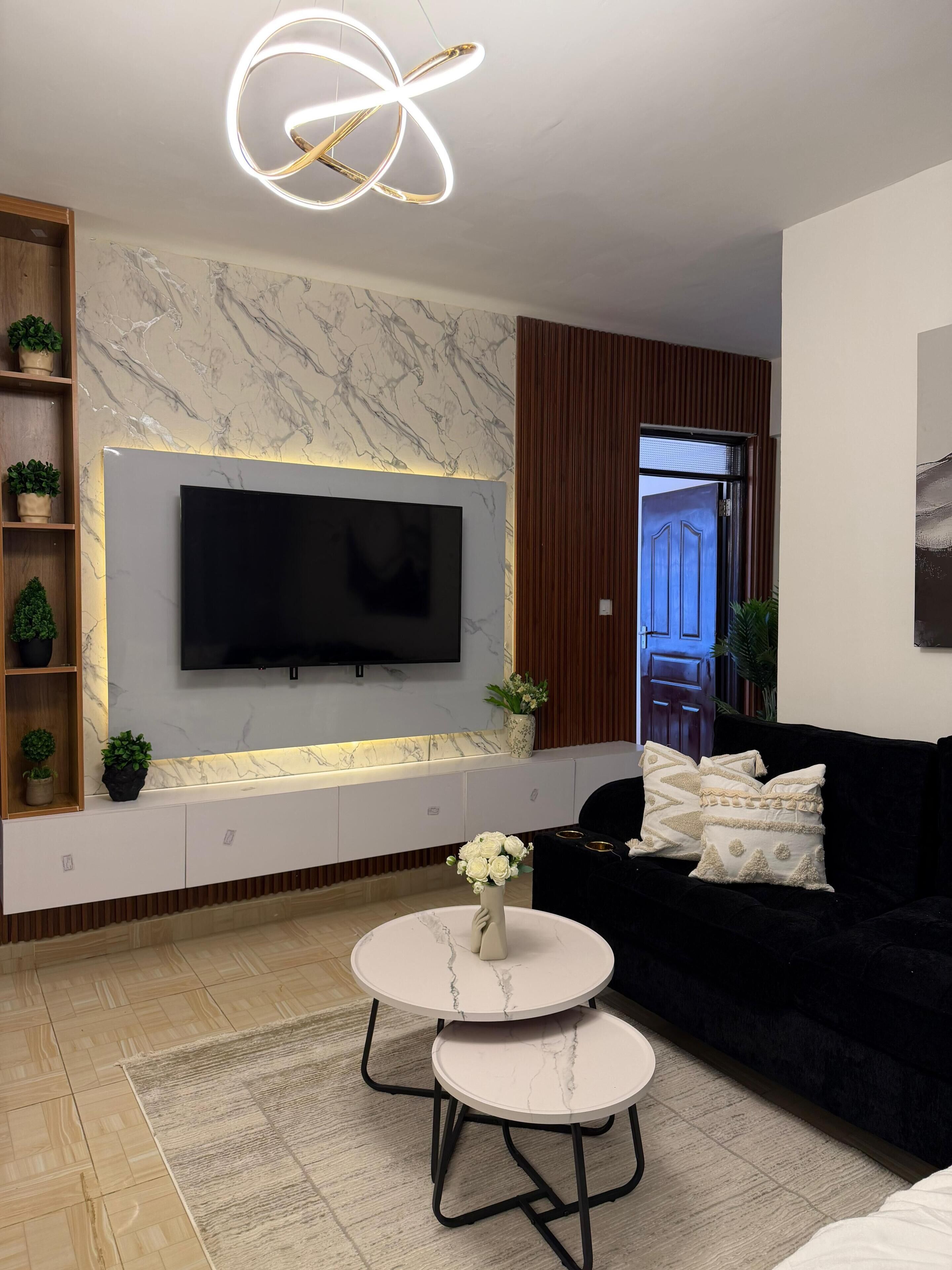 Ein modernes Wohnzimmer mit Flachbildfernseher an einer Marmorwand, einer schwarzen Couch, einem weißen Couchtisch und einer außergewöhnlichen Deckenleuchte.