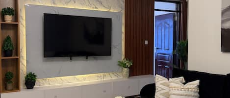 A modern living room with a flat-screen TV mounted on a marble wall, a black sofa, a white coffee table, and a unique ceiling light fixture.