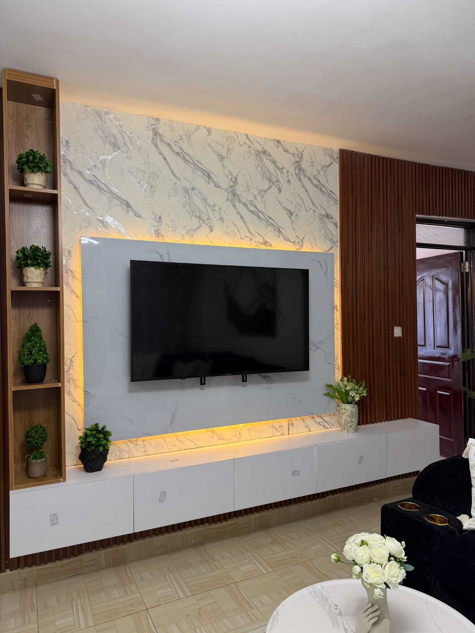 Un salon moderne doté d’un grand téléviseur à écran plat fixé sur un mur en marbre, d’étagères en bois ornées de plantes en pot et d’un canapé de couleur foncée.