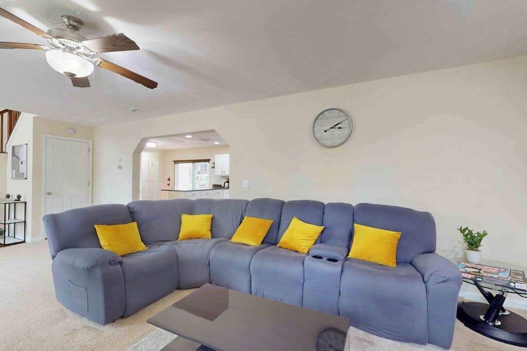 Ein Wohnzimmer mit grauem Ecksofa, einem Couchtisch, einem Deckenventilator und einer Wanduhr.