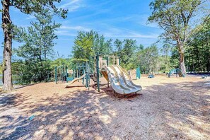 Ein Spielplatz mit Rutsche und Klettergerüsten, umgeben von Bäumen und einem klaren Himmel.