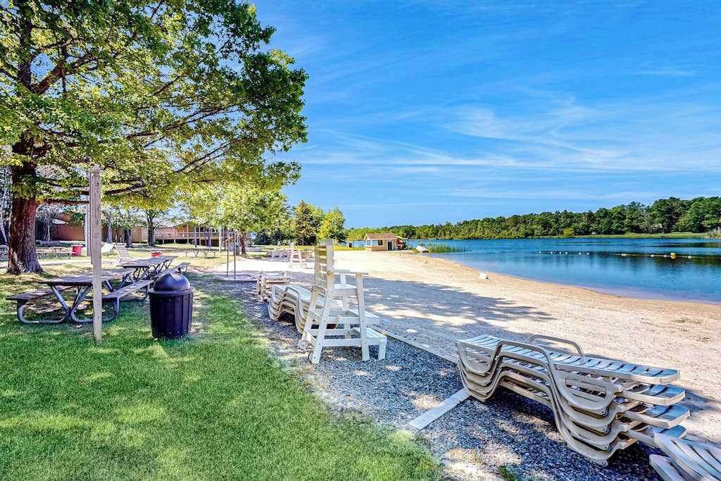 A lakeside park with picnic tables, lounge chairs, and a trash bin.