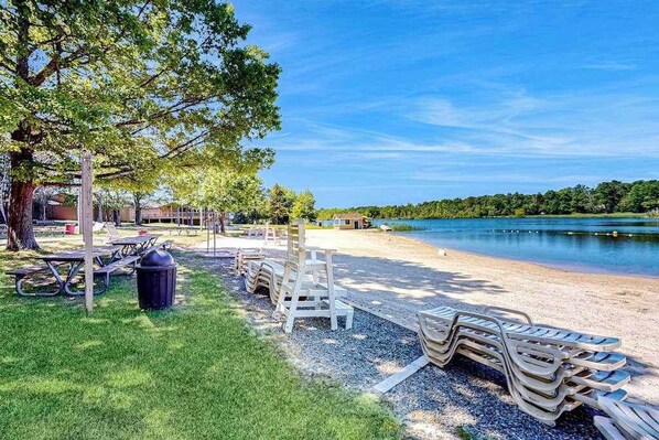 A lakeside park with picnic tables, lounge chairs, and a trash bin. - Pocono Home with Hot Tub, EV Charge, Pool & Tennis (Tobyhanna)