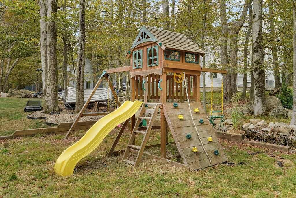 A wooden play structure with a slide and climbing wall, surrounded by trees.