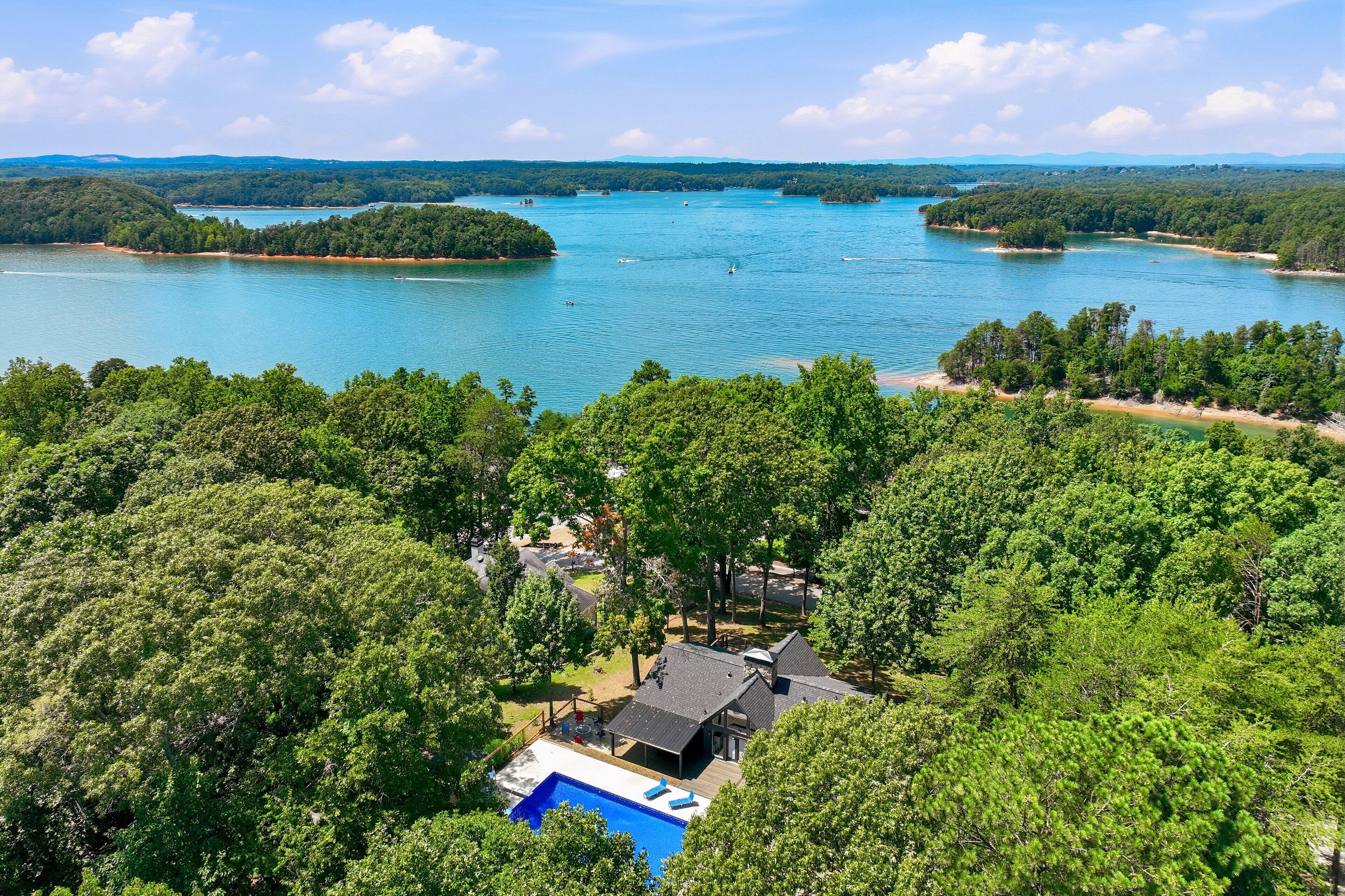 A residential area with a swimming pool, surrounded by dense forest and a large body of water with boats.