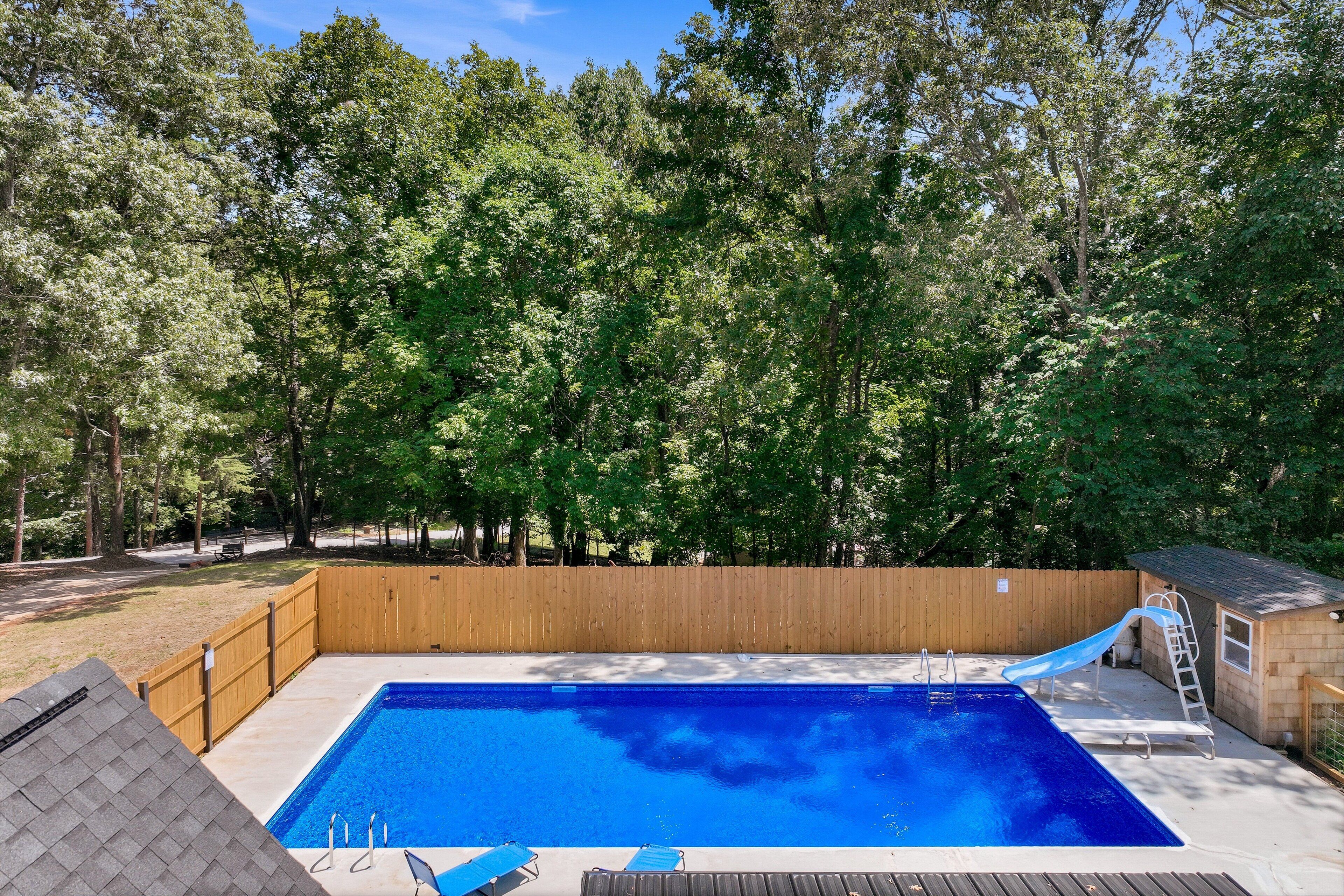Ein rechteckiger Pool mit Holzterrasse und Rutsche, umgeben von einem Holzzaun und Bäumen.