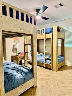 Ein Zimmer mit Etagenbetten, einem Deckenventilator, einem kleinen Tisch mit Lampe und einem Fenster mit Jalousien.