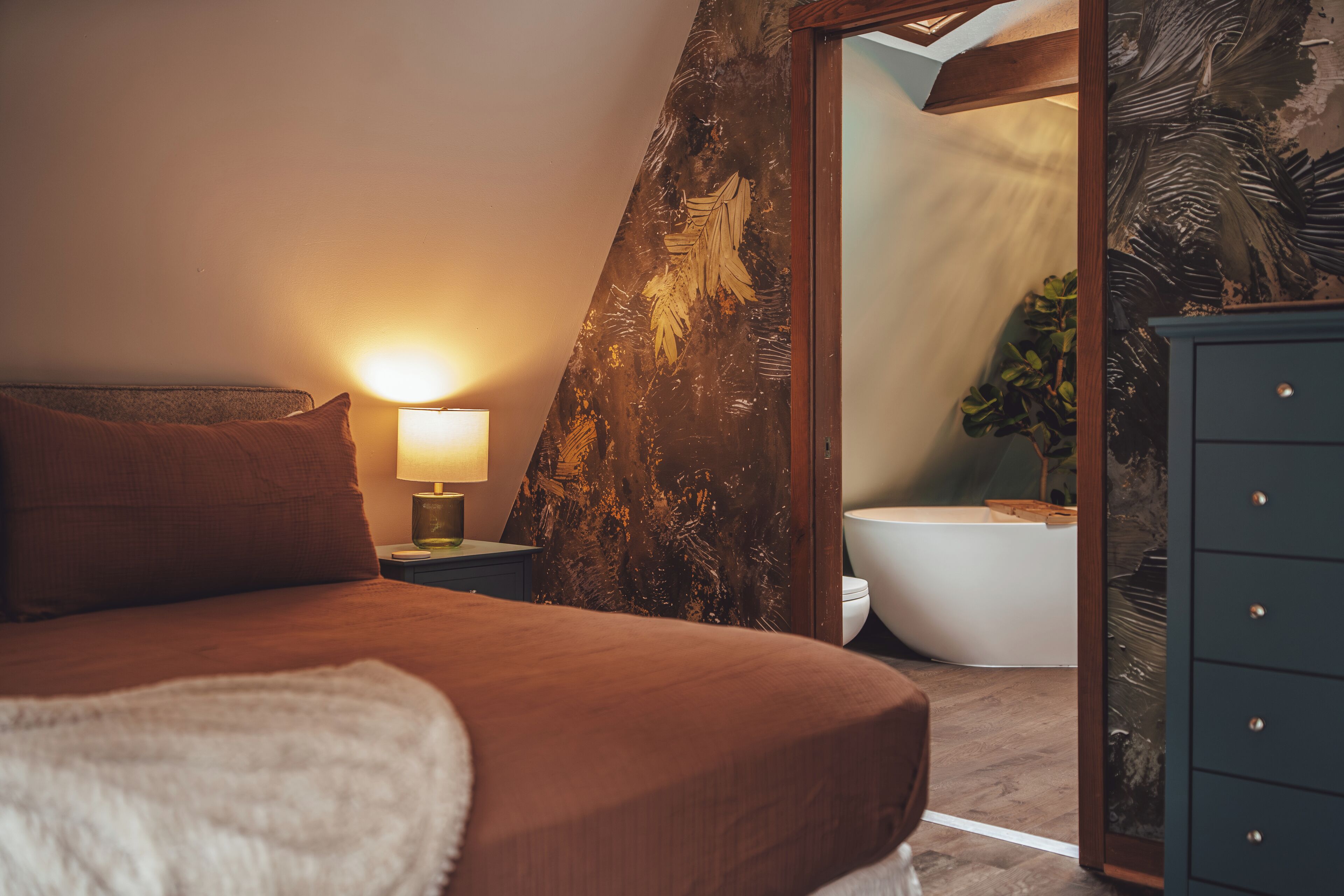 Une chambre à coucher avec un lit, une lampe de chevet et une vue sur la salle de bain à travers une porte.