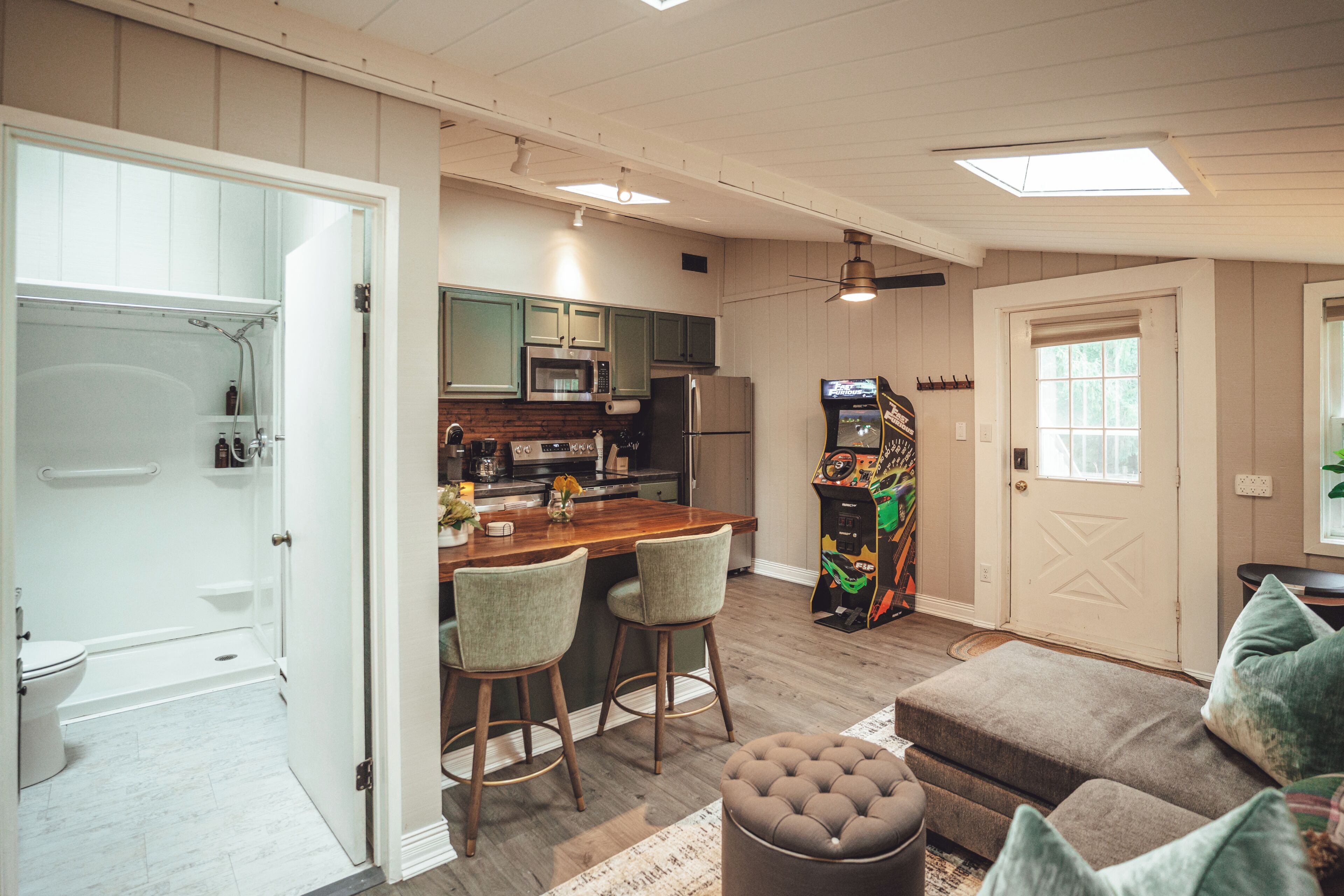 Un espace de vie compact comprenant un coin cuisine, un espace repas avec des tabourets de bar, une console de jeux et un coin salon confortable avec un canapé et un pouf.