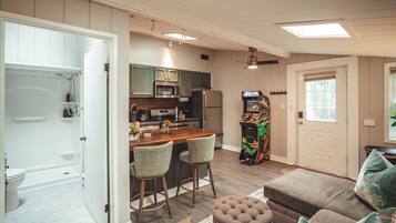 Un espace de vie compact comprenant un coin cuisine, un espace repas avec des tabourets de bar, une console de jeux et un coin salon confortable avec un canapé et un pouf.