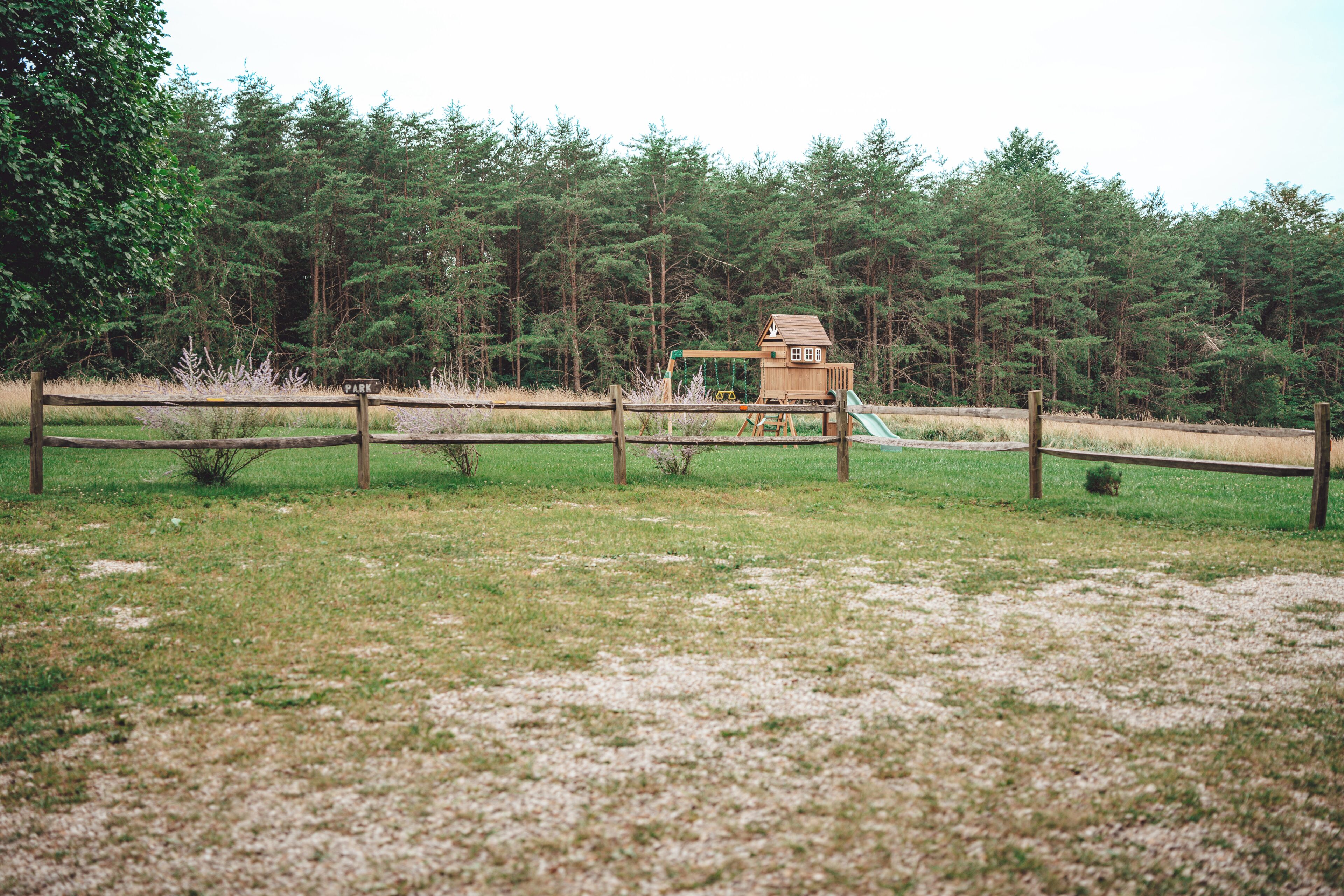 Una casita de juegos de madera con un tobogán en un campo cubierto de césped, rodeada de una valla de madera y árboles frondosos al fondo.