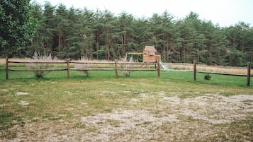 Una casita de juegos de madera con un tobogán en un campo cubierto de césped, rodeada de una valla de madera y árboles frondosos al fondo.
