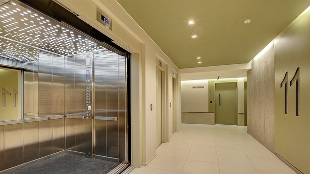 Um elevador moderno com acabamento metálico e painel de comandos, que dá acesso a um corredor com chão em azulejos e uma porta com o número 11.