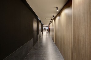 Ein langer, schmaler Flur mit Türen an einer Seite und einer Holzwand an der anderen, der zu einem Raum mit einem Sessel und einem Tisch führt.