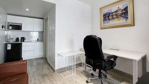 Quarto moderno e minimalista com escrivaninha branca, cadeira de escritório preta, sofá de couro marrom e área de cozinha com armários brancos e geladeira preta.