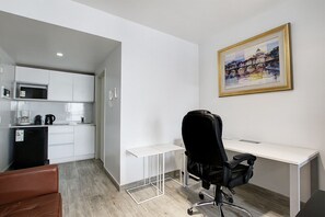 Ein modernes, minimalistisches Zimmer mit einem weißen Schreibtisch, einem schwarzen Bürostuhl, einer braunen Ledersofa und einer Küchenzeile mit weißen Schränken und einem schwarzen Kühlschrank.