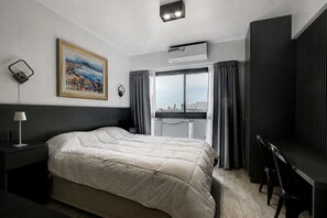 Ein Hotelzimmer mit einem Bett, einem Schreibtisch, einem Stuhl, einem Fenster mit Vorhängen, einer Klimaanlage und einem Bild an der Wand.