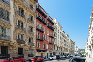 A row of multi-story buildings with balconies, a street with parked cars, and storefronts with signage. - Appartement Le Verdi avec balcon  - Welkeys (Nice)