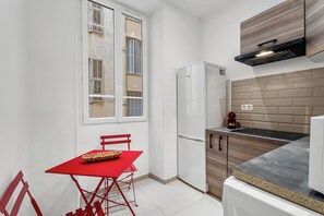 A compact kitchen with a refrigerator, a built-in oven, and a window with shutters. - Appartement Le Verdi avec balcon  - Welkeys (Nice)