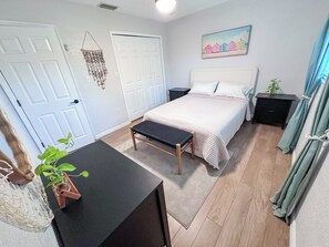 A bedroom with a bed, bedside tables, a desk, a hanging plant, and a painting on the wall.
