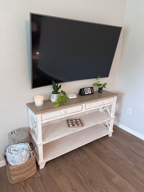 A living room with a flat-screen TV mounted on the wall, a wooden console table with a candle, a small potted plant, and a calendar.