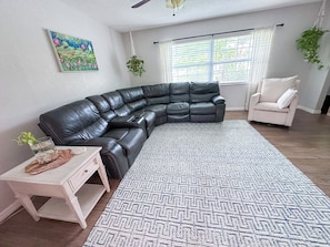 A living room with a sectional sofa, a small table with a plant, a patterned rug, and a window with blinds.
