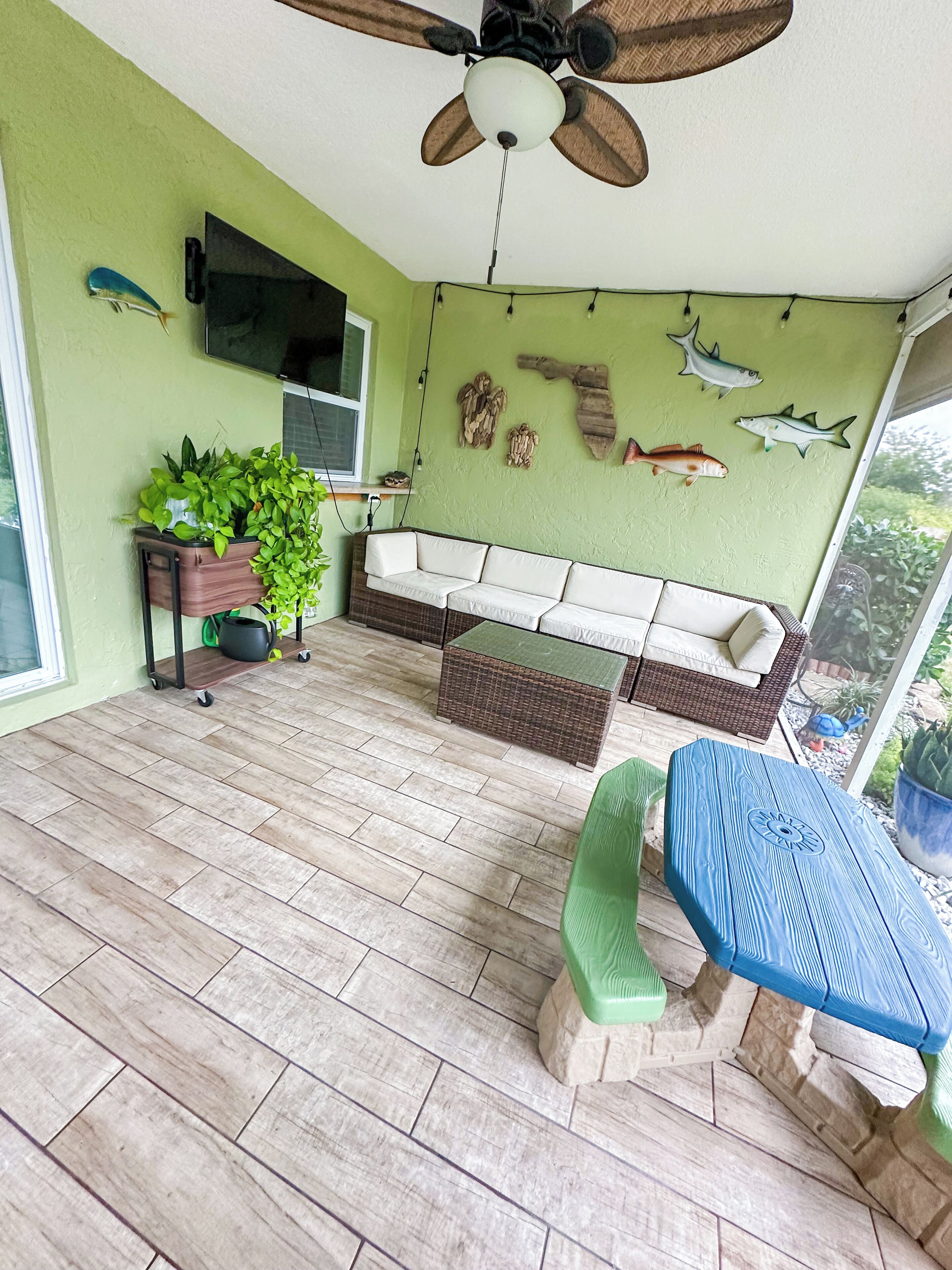 A room with a ceiling fan, a white sofa, a green bench, a blue table, and a wall with fish decorations.