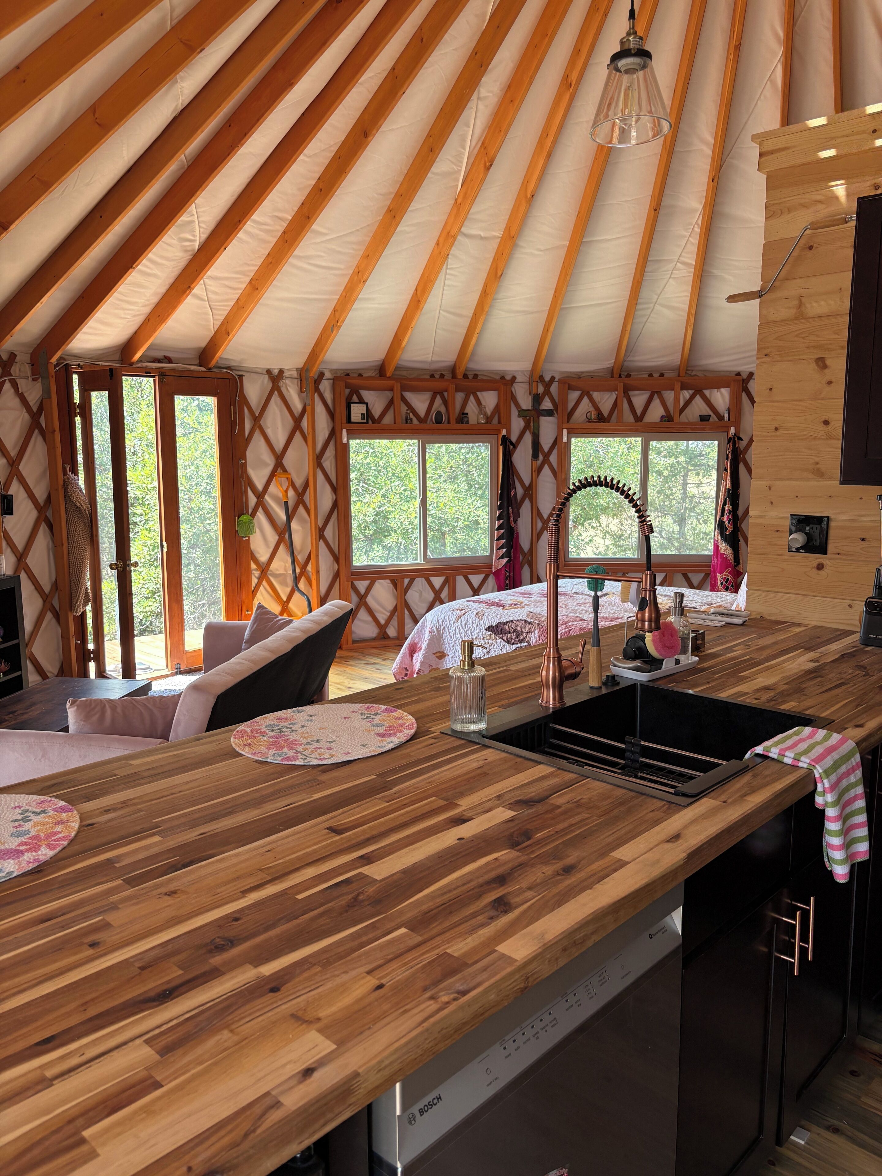A yurt interior with a wooden dining table, kitchen area, and a large window providing a view of the outdoors.