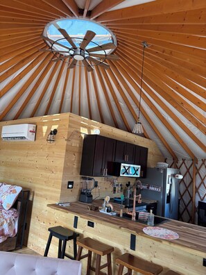 Interior d'una iurta amb sostre de fusta, un ventilador central, una zona de cuina amb nevera i un tamboret.