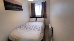 A small, single-bed room with a painting on the wall, a window with curtains, and a radiator. - Joydens Wood, 2-Bed Apt, Sleeps 4 with Parking (Essex)
