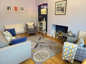 A living room with a sofa, armchair, and a coffee table. There are framed pictures on the wall, a bookshelf, and a fireplace. - Largo Bay - Harbour Hideaway (Leven)
