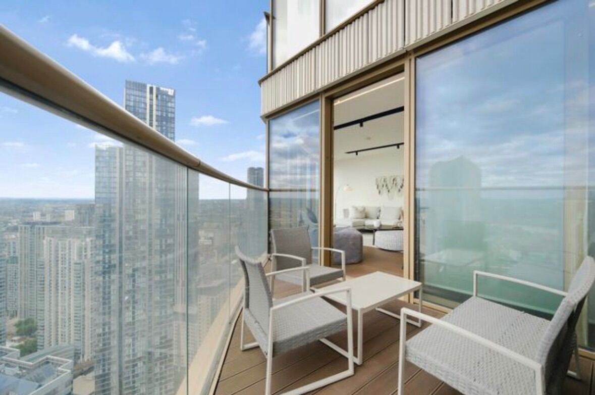 Een balkon op een hoog gebouw met een glazen hekwerk, voorzien van witte stoelen en een kleine tafel, met uitzicht op een stadsgezicht.