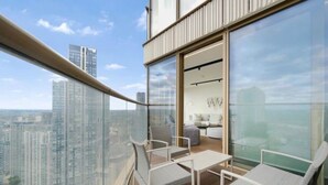 Ein hoher Balkon mit Glasgeländer, ausgestattet mit weißen Stühlen und einem kleinen Tisch, mit Blick auf eine Stadtlandschaft.