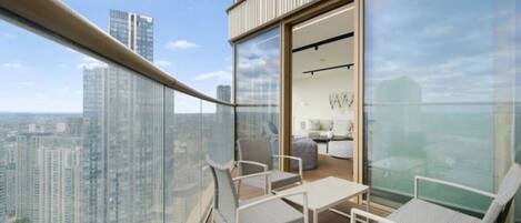 A high-rise balcony with a glass railing, furnished with white chairs and a small table, overlooking a cityscape.