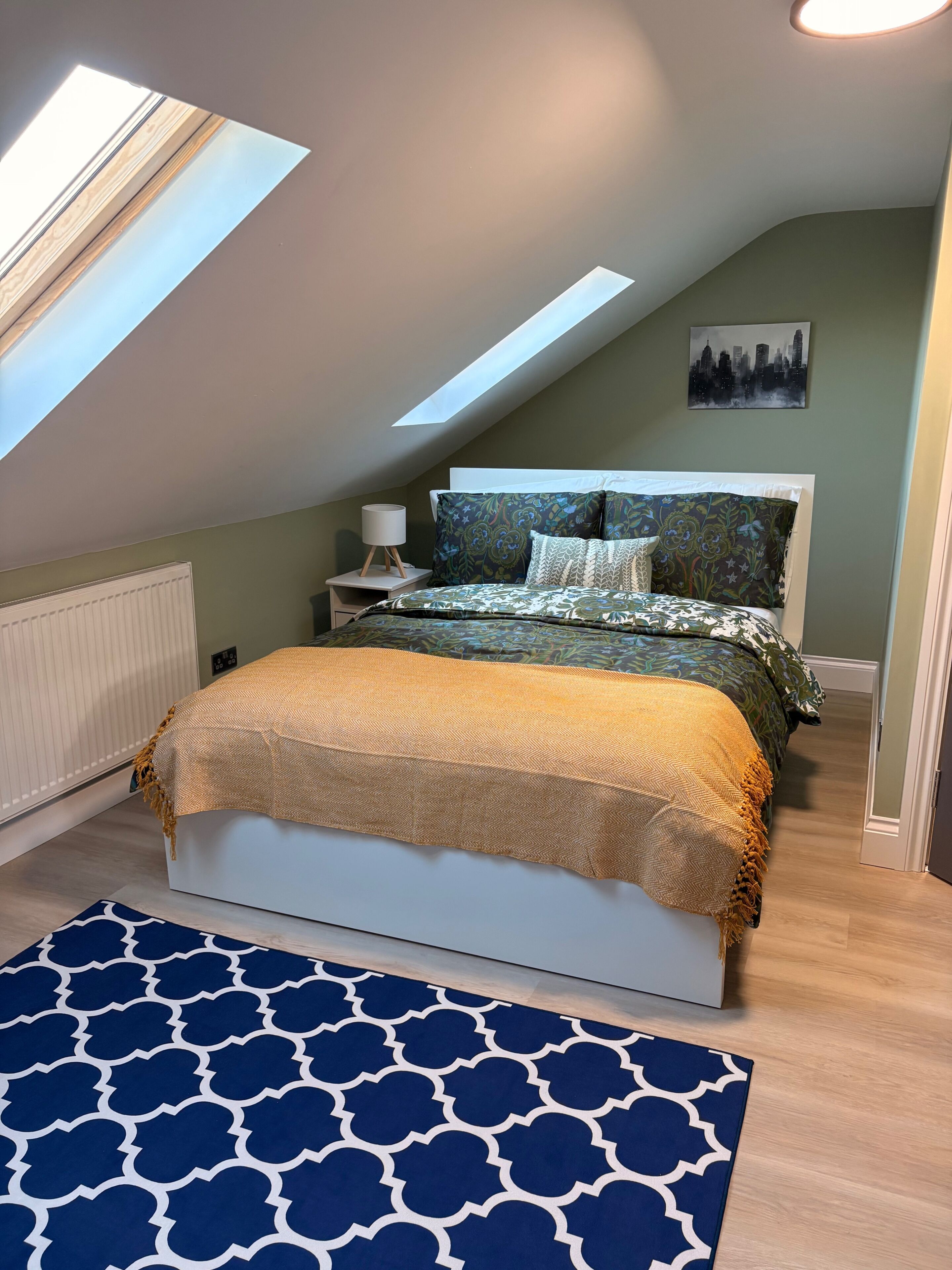Une chambre avec une lucarne, un lit recouvert d’une literie à motifs, une table de chevet avec une lampe et un tableau représentant un paysage urbain accroché au mur.