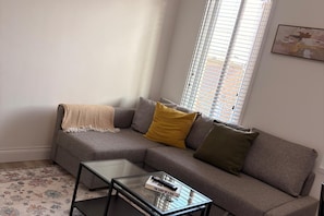 Una sala de estar con un sofá gris, una mesa de centro de vidrio y un ventanal con persianas.