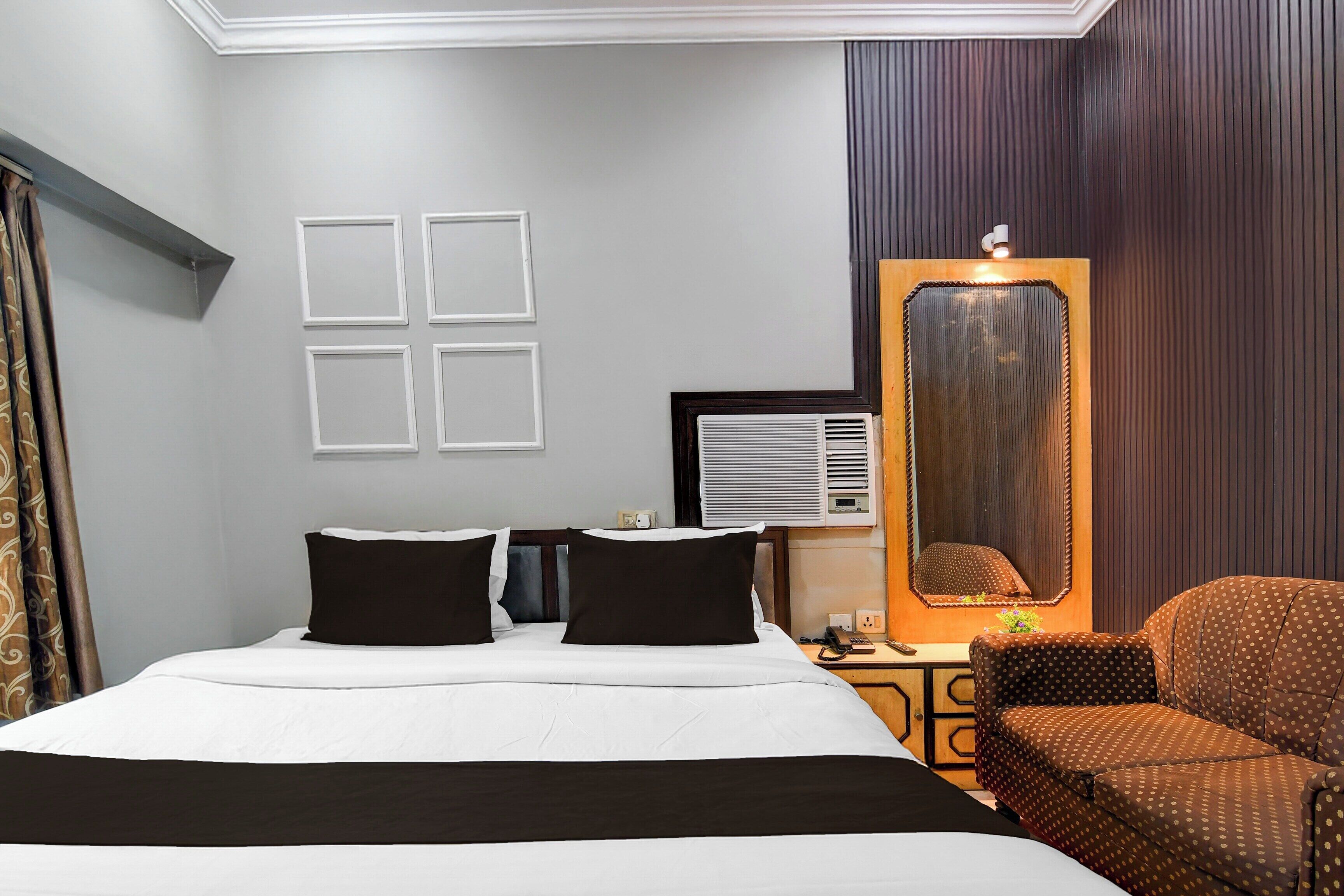 A hotel room with a bed, a chair, a mirror, and a wall-mounted air conditioning unit.