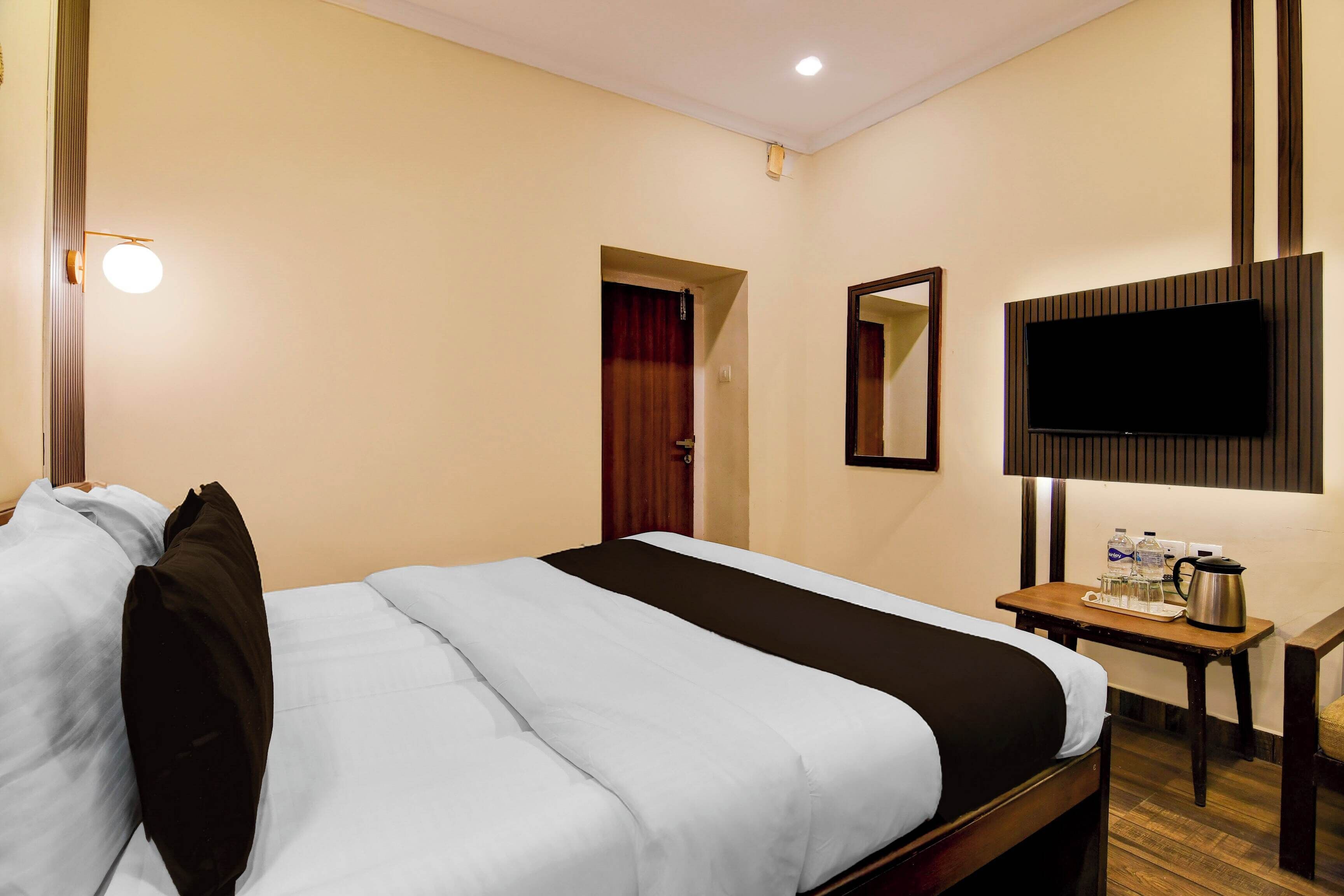 Habitación de hotel con cama, televisor montado en la pared, una mesita con hervidor y botellas de agua, y una puerta que da a otra habitación.