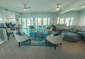 A spacious living room with a large blue rug, white armchairs, a sofa, and a coffee table. There are ceiling fans, a dining area with a table and chairs, and large windows offering a view of the outdoors. - Beautiful Home in Port Antigua with Dock and Private Beach Access (Islamorada)