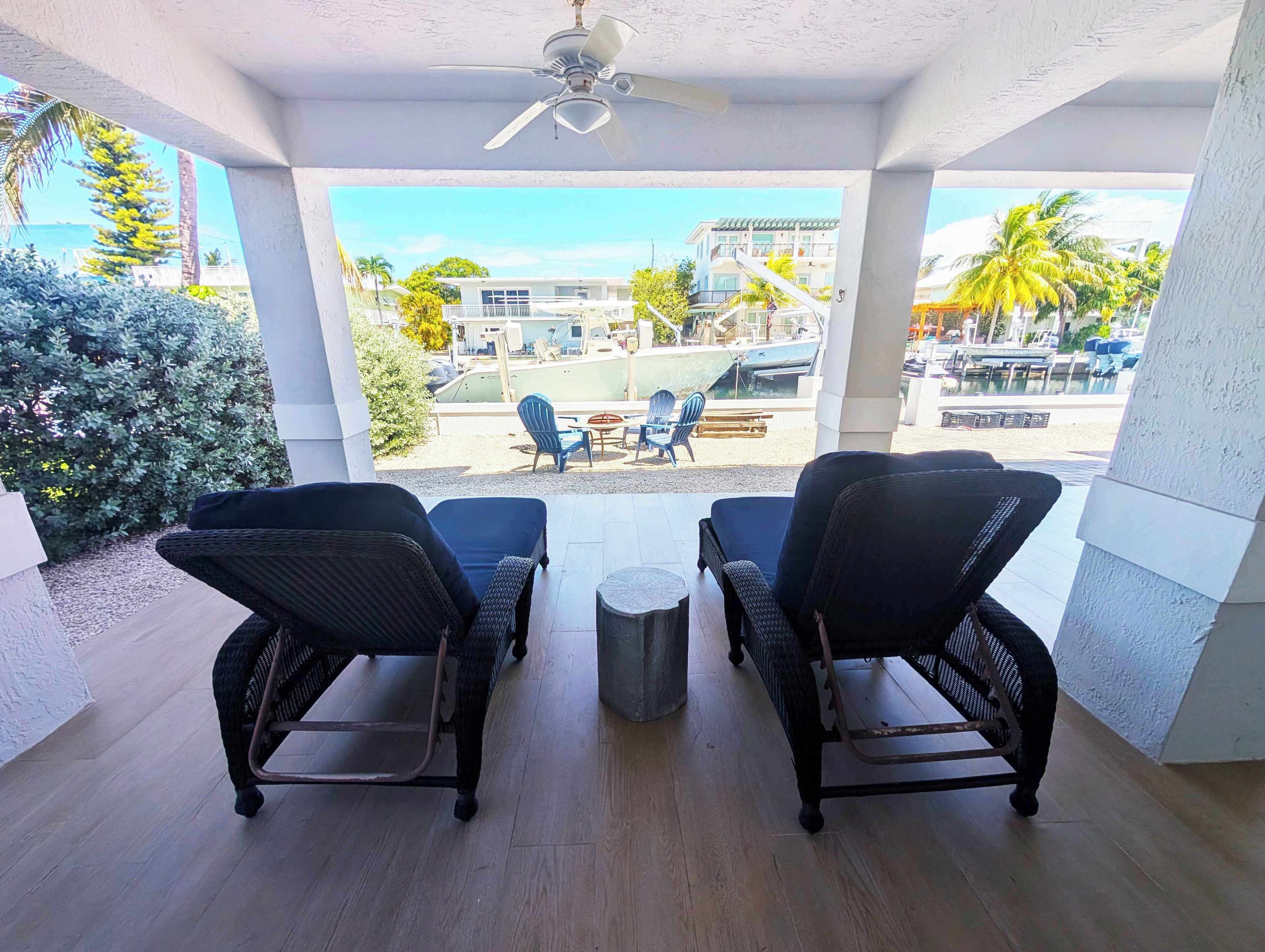 A covered outdoor seating area with two wicker chairs and a small table, overlooking a marina with boats and buildings.