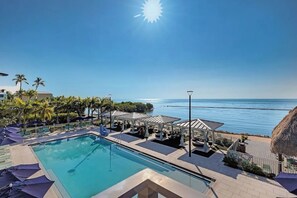 Ein Poolbereich mit Meerblick, Liegestühle, Sonnenschirme und ein strohgedecktes Gebäude.