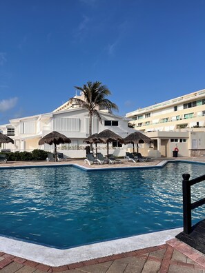 Ein Swimmingpool mit Liegestühlen und Sonnenschirmen, eine Palme und ein Gebäude mit Balkonen.
