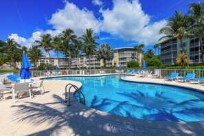 Ein Pool mit Liegestühlen und Sonnenschirmen, umgeben von Palmen und einem Hotelgebäude.